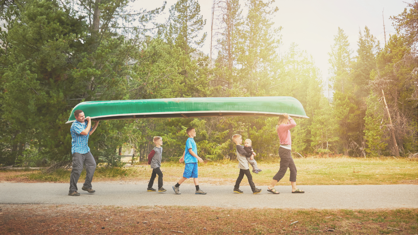 A family with four children carrying a canoe to the water near their RV campsite, enjoying a casual camping adventure.